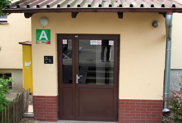 Wejście do budynku z brązowymi drzwiami i domofonem. Nad drzwiami znak zakazu palenia, po lewej stronie zielony szyld z literą A i numerem. Rynna po prawej.