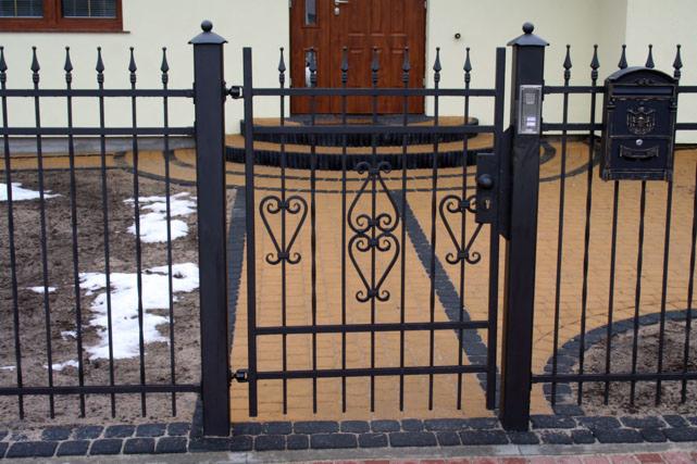 Czarna, metalowa brama z dekoracyjnymi elementami w kształcie serc i domofonem, skrzynka na listy na tle domu z brązowymi drzwiami i chodnikiem z kostki.