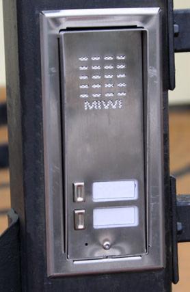 Metalowy panel wejściowy z przyciskami i miejscem na nazwę firmy 'MIWI', zamontowany na czarnym, metalowym ogrodzeniu posesji.