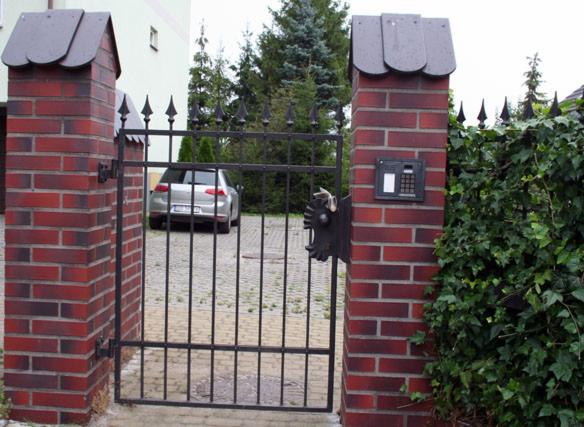 Brama wjazdowa z ceglanymi słupkami i domofonem, ozdobnym elementem metalowym w kształcie głowy lwa, widok na podjazd i samochód.