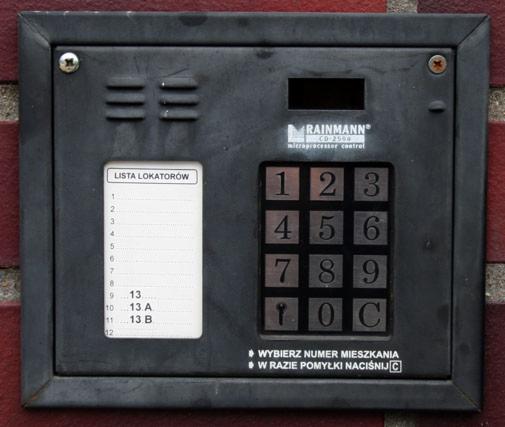 Czarny panel domofonowy Rainmann CD-2564 z listą lokatorów (1-13B) i klawiaturą numeryczną, zamontowany na ceglanej ścianie. Widoczne śruby mocujące.