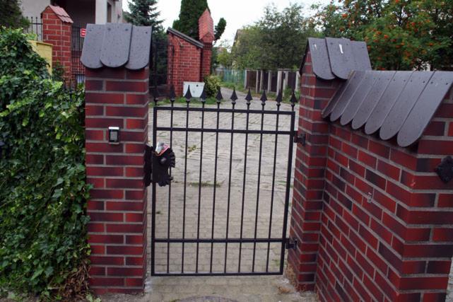 Kuta, czarna furtka z domofonem na ceglanym słupku z daszkiem. Widok z zewnątrz posesji, w tle zabudowa jednorodzinna i zieleń. Klasyczny styl.