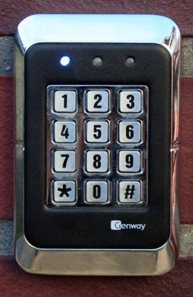 Czarny panel numeryczny Genway z chromowaną ramką, zamontowany na ceglanej ścianie. Drobne przyciski z cyframi i symbolami. Świecąca niebieska dioda.