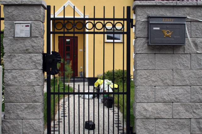 Czarna, metalowa furtka z ozdobnymi elementami, zamontowana między dwoma szarymi, kamiennymi słupkami. Na jednym ze słupków zamontowany domofon i skrzynka na listy.