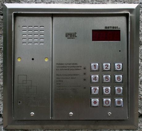 Panel wejściowy z klawiaturą numeryczną i wyświetlaczem LED, marki Urmet i Matibus, zamontowany na szarej ścianie. Widoczne instrukcje obsługi i przyciski wyboru numeru lokalu.