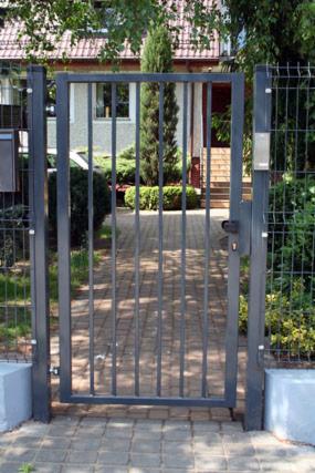 Metalowa furtka z zamkiem i domofonem przy posesji. Widok na dom z ogrodem przez pręty furtki. Szara kostka brukowa na podjeździe.