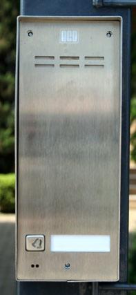 Stalowy panel domofonu z przyciskiem dzwonka i miejscem na nazwisko, zamontowany na czarnym metalowym słupku bramy na tle zieleni.