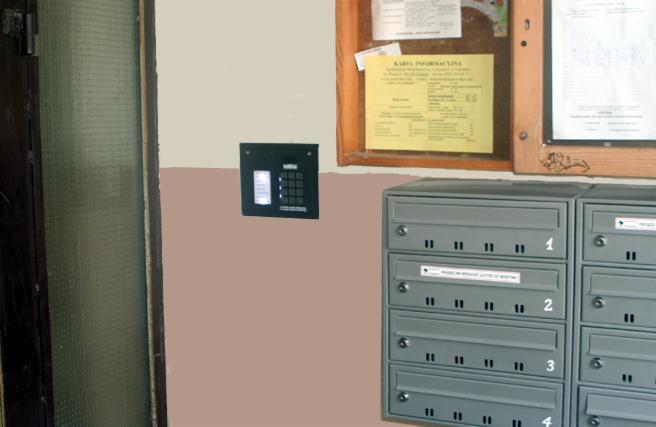 Zamontowany czarny panel z klawiaturą numeryczną obok szarych skrzynek pocztowych w bloku. Tablica informacyjna nad skrzynkami.