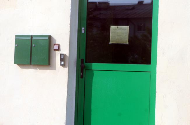 Zielone skrzynki pocztowe i panel  przy wejściu z  domofonem na białej ścianie obok zielonych drzwi z szybą, na której wisi kartka.