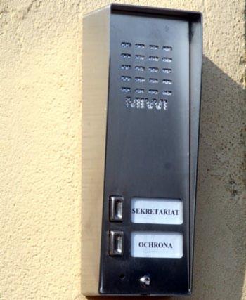 Elegancki, czarny panel z przyciskami 'Sekretariat' i 'Ochrona' zamontowany na beżowej ścianie budynku. Widoczny szyfrator i głośnik.