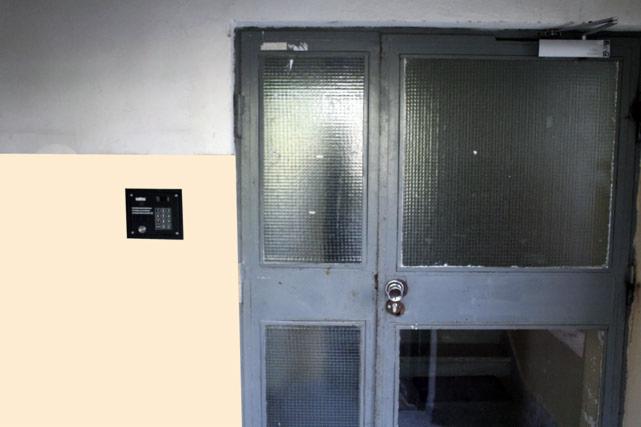 Szary panel domofonu z klawiaturą numeryczną zamontowany na ścianie obok szklanych drzwi wejściowych do budynku.