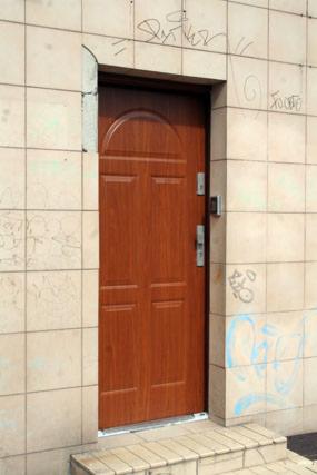 Brązowe drzwi wejściowe z domofonem w bloku z beżową elewacją. Widoczne ślady graffiti na ścianie.