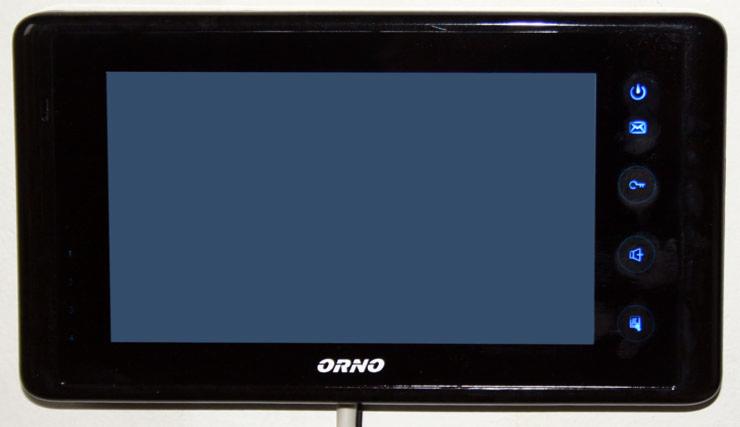 Czarny wideodomofon ORNO z niebieskimi ikonami funkcyjnymi po prawej stronie i szarym ekranem, zamontowany na białej ścianie.