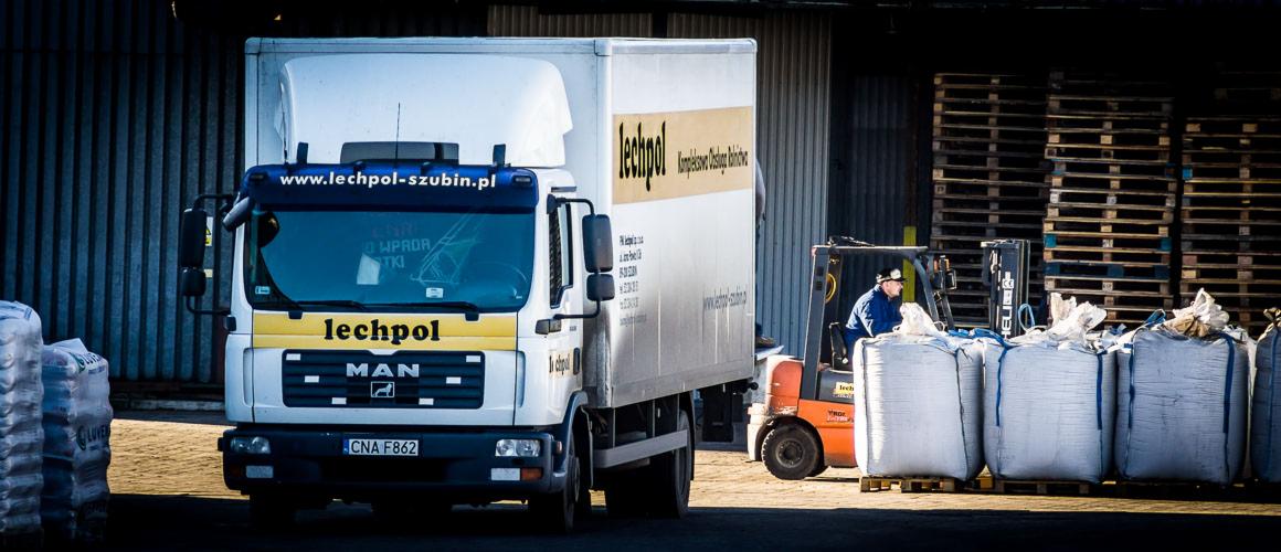 Ciężarówka MAN firmy Lechpol, w tle wózek widłowy i palety, sugerujące załadunek lub rozładunek towarów. Widoczne logo firmy na pojeździe.
