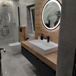 MG MEBLE MICHAŁ GRYT - Nowoczesna łazienka z drewnianymi akcentami: szafka pod umywalkę z czarnymi frontami, prostokątna umywalka nablatowa, okrągłe lustro z podświetleniem LED, toaleta podwieszana i kabina prysznicowa...