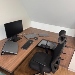MG MEBLE MICHAŁ GRYT - Ergonomiczne biurko narożne z drewna orzechowego z wbudowanymi szafkami, czarny monitor, laptopy i klawiatura, krzesło biurowe z regulowanym zagłówkiem.
