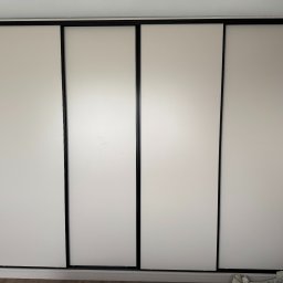 MG MEBLE MICHAŁ GRYT - Czterodrzwiowa szafa wnękowa z przesuwnymi, białymi frontami i czarnymi ramami, widoczna z boku drzwi wejściowe i fragment ściany z wykończeniem drewnopodobnym.