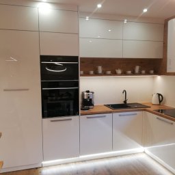 MG MEBLE MICHAŁ GRYT - Nowoczesna kuchnia z białymi, lakierowanymi frontami, drewnianym blatem i podświetleniem LED w dolnej części szafek. Na półce dekoracyjne filiżanki i imbryk.