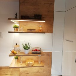 MG MEBLE MICHAŁ GRYT - Półki z podświetleniem LED w nowoczesnej kuchni, dekoracyjne przedmioty i produkty spożywcze na drewnianych półkach, białe fronty szafek w tle.