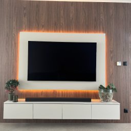 MG MEBLE MICHAŁ GRYT - Nowoczesna ściana TV z fornirowaną okładziną, podświetleniem LED, białą szafką RTV, soundbarem i dekoracyjnymi roślinami w doniczkach.