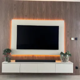 Nowoczesna ściana TV z fornirowaną okładziną, podświetleniem LED, białą szafką RTV, soundbarem i dekoracyjnymi roślinami w doniczkach.