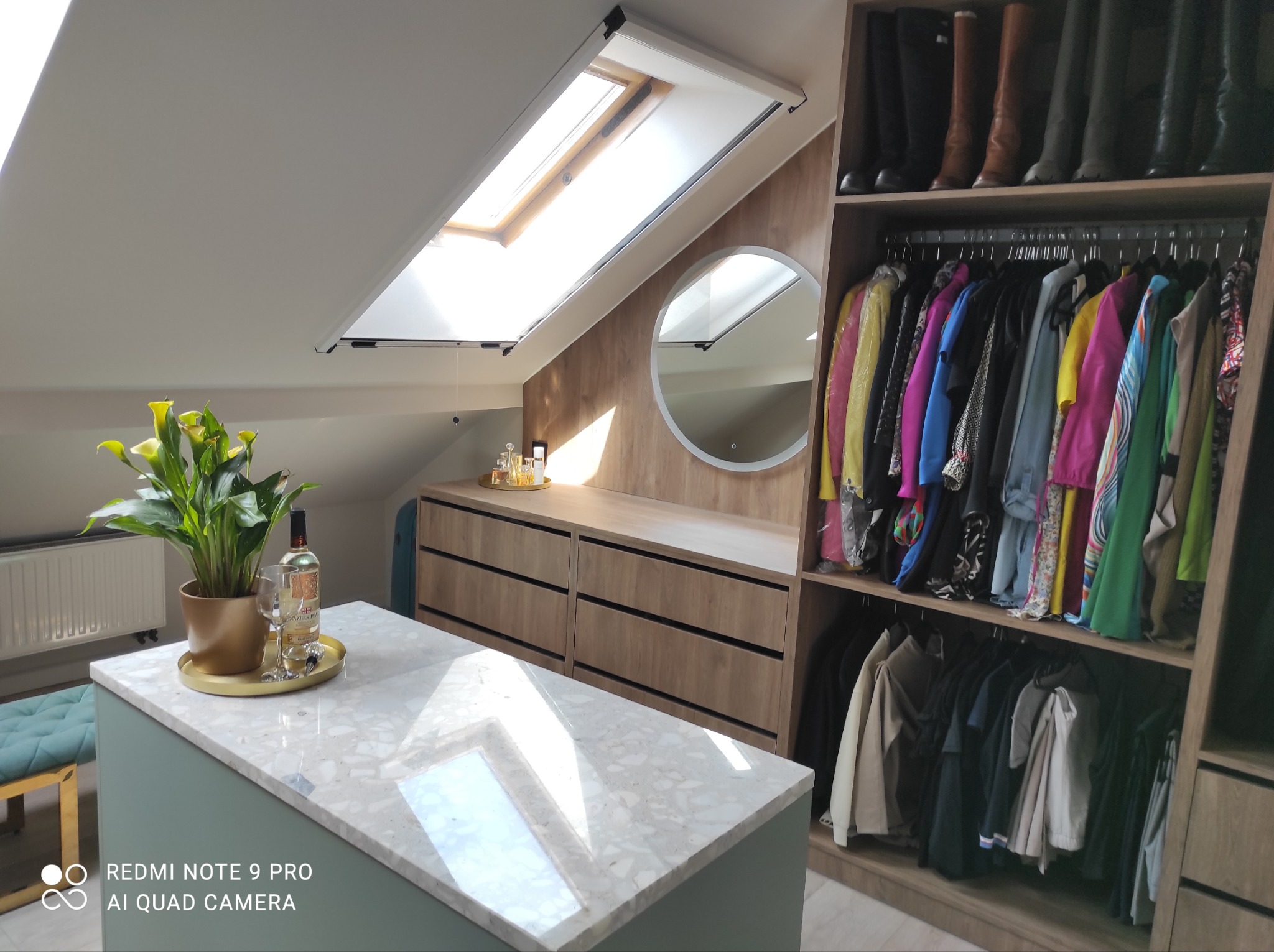 Garderoba na poddaszu z oknem dachowym: szafa wnękowa z ubraniami i butami, komoda z lustrem i dekoracjami, wyspa z blatem imitującym marmur i roślinami w doniczce.