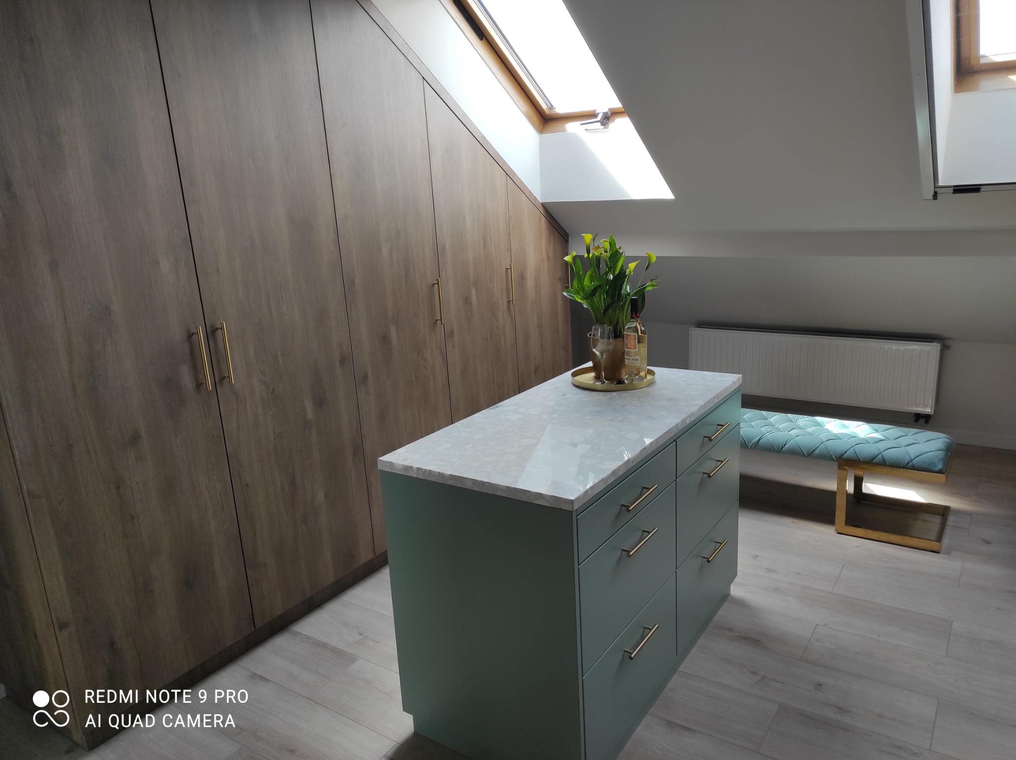 Szafa wnękowa z drewnianymi frontami i wyspa kuchenna z blatem imitującym marmur, złote uchwyty, poddasze z oknem dachowym.