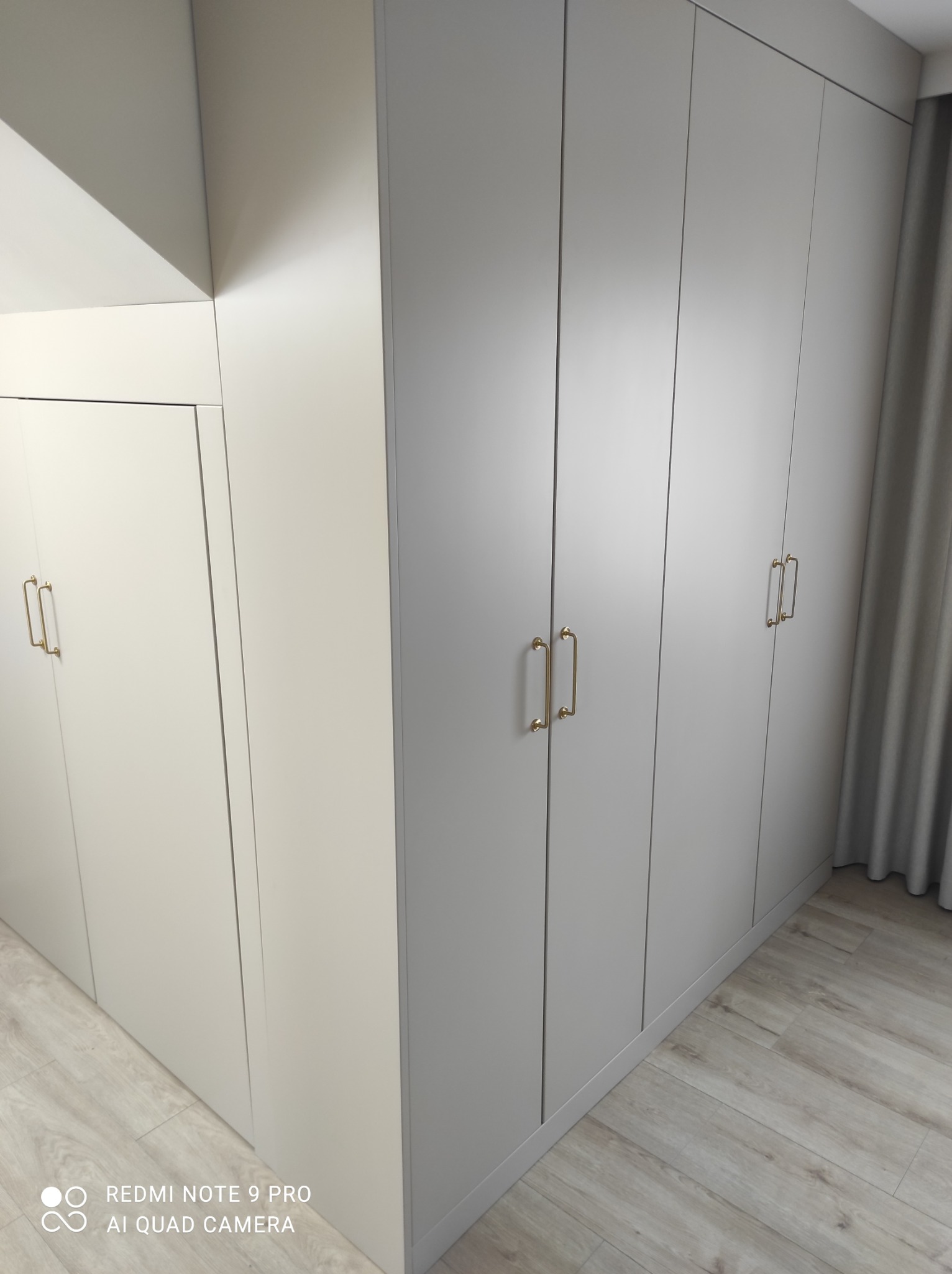 Jasnoszara zabudowa szafy wnękowej z złotymi uchwytami, widoczna część podłogi z paneli oraz zasłona w tle