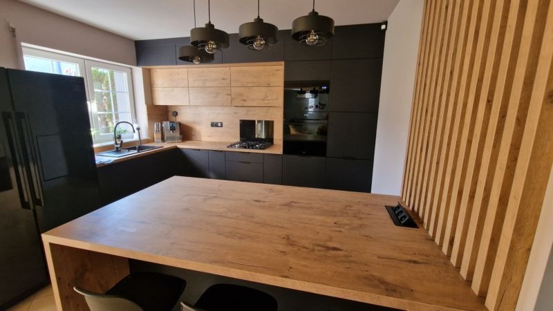 Nowoczesna kuchnia z czarnymi szafkami i drewnianymi frontami, wyspa kuchenna z drewnianym blatem i ukrytymi gniazdkami, dekoracyjna ściana z drewnianych listew.