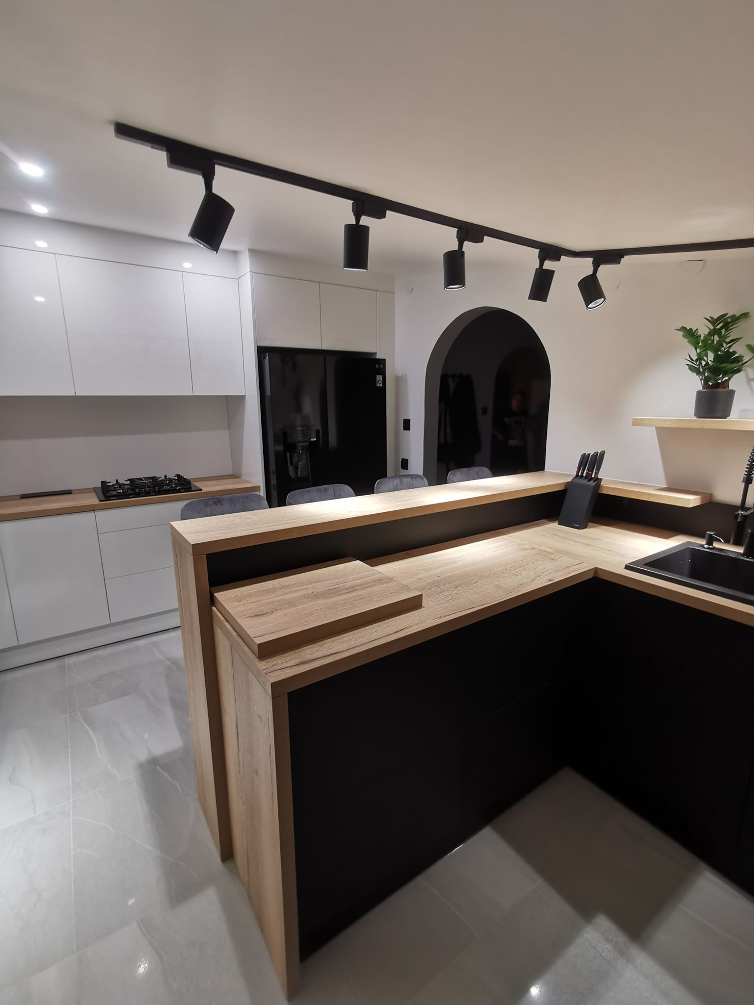 Nowoczesna kuchnia z wyspą, czarne fronty szafek dolnych i drewniany blat, oświetlenie punktowe na szynie, płyta grzewcza na blacie, czarna lodówka, podłoga z jasnych płytek.