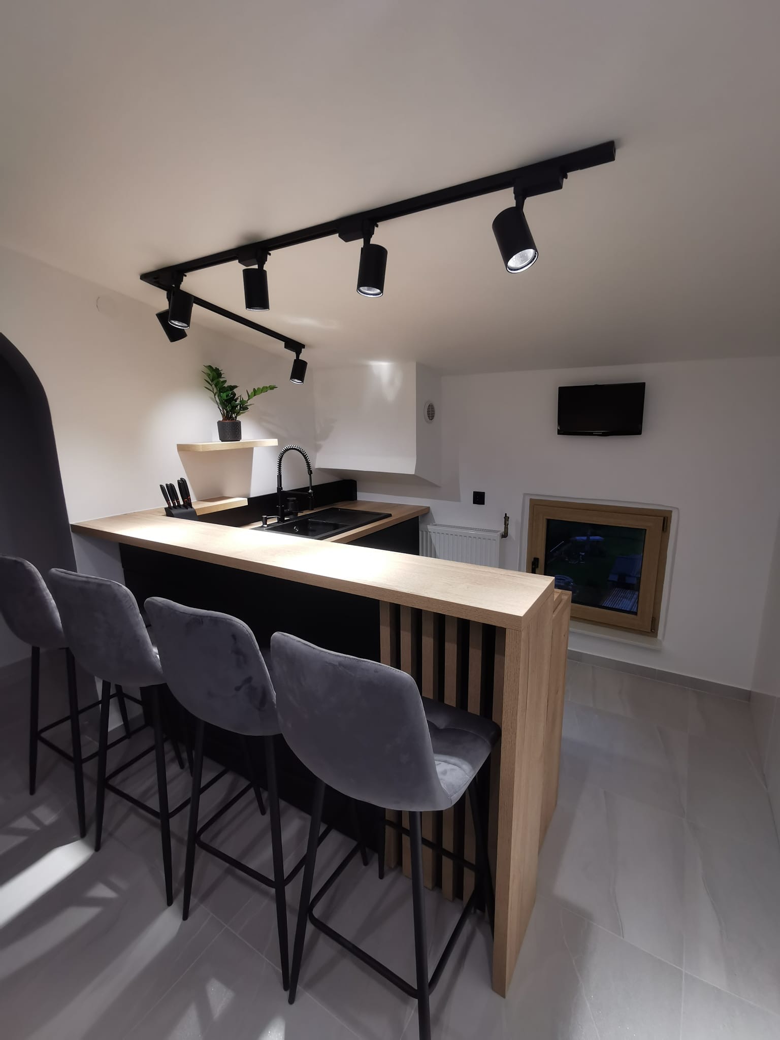Nowoczesna kuchnia z czarnym blatem, drewnianym barkiem z pionowymi listwami, szarymi hokerami i czarną armaturą, oświetlona reflektorami szynowymi.