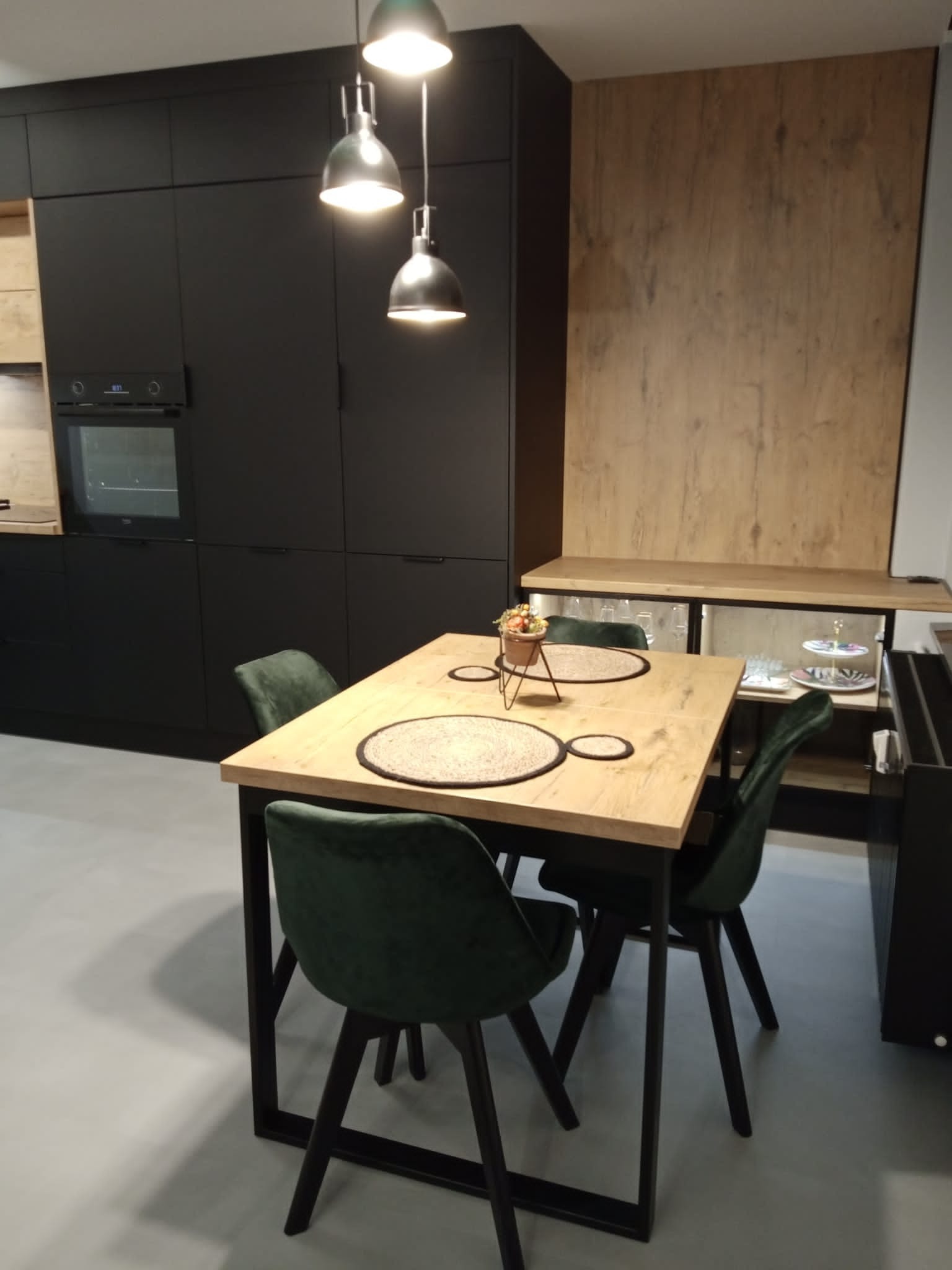 Nowoczesna jadalnia z czarnymi szafkami kuchennymi, drewnianym blatem stołu i zielonymi krzesłami, oświetlona trzema wiszącymi lampami.