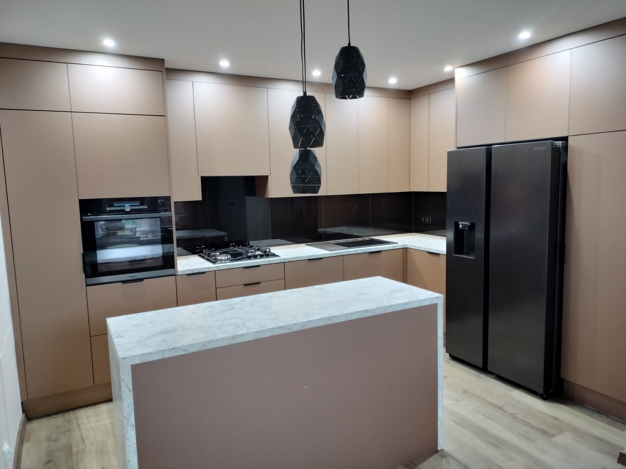 Nowoczesna kuchnia z beżowymi frontami, wyspą z marmurowym blatem i czarną lodówką side-by-side, oświetlona trzema wiszącymi lampami o geometrycznym kształcie.