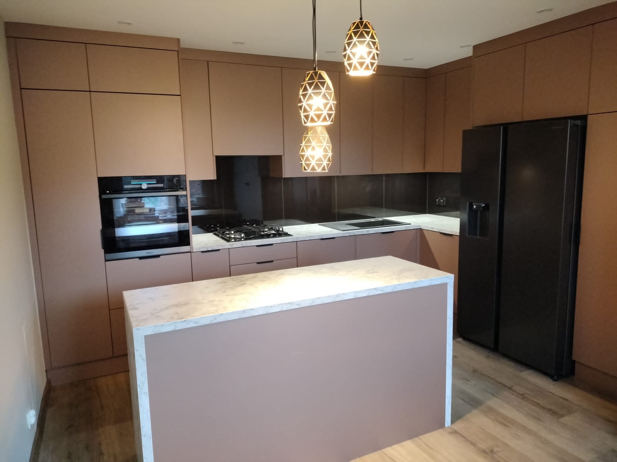 Nowoczesna kuchnia z zabudową w kolorze pudrowego różu, wyspa z marmurowym blatem, czarna lodówka side-by-side i dekoracyjne lampy wiszące.