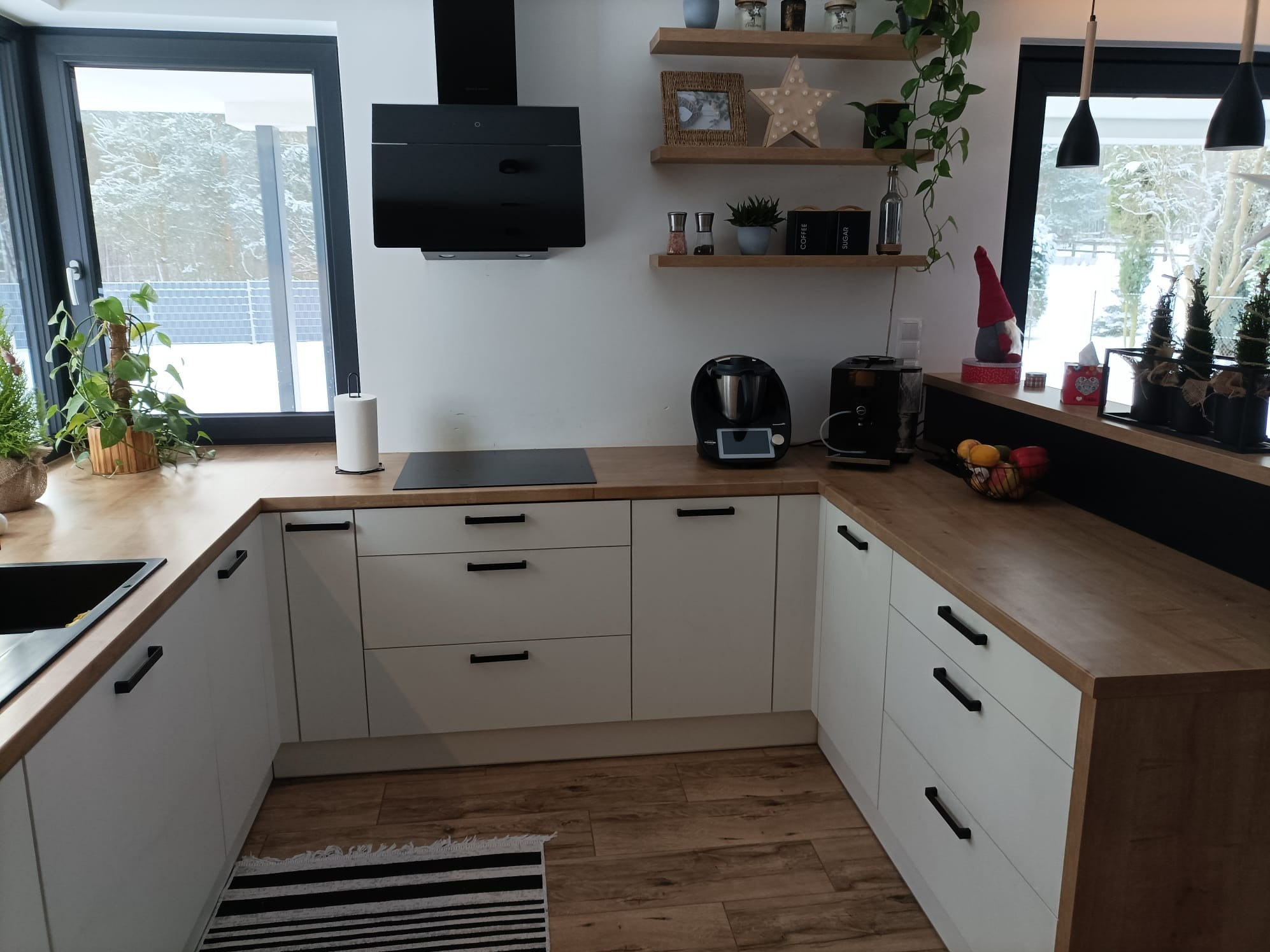 Nowoczesna, jasna kuchnia z białymi frontami szafek i drewnianym blatem, z czarnymi uchwytami, okapem i zlewem, ozdobiona roślinami i dekoracjami, widok z perspektywy podłogi.