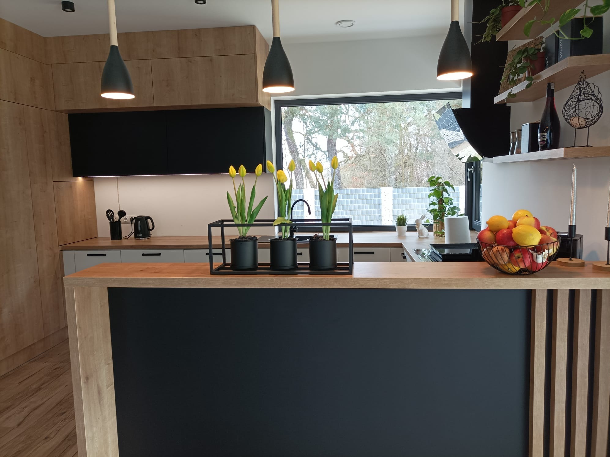 Nowoczesna kuchnia z drewnianymi frontami i czarnymi akcentami, wyspa kuchenna z czarnym panelem i dekoracją z tulipanów, oświetlenie punktowe i wiszące lampy.
