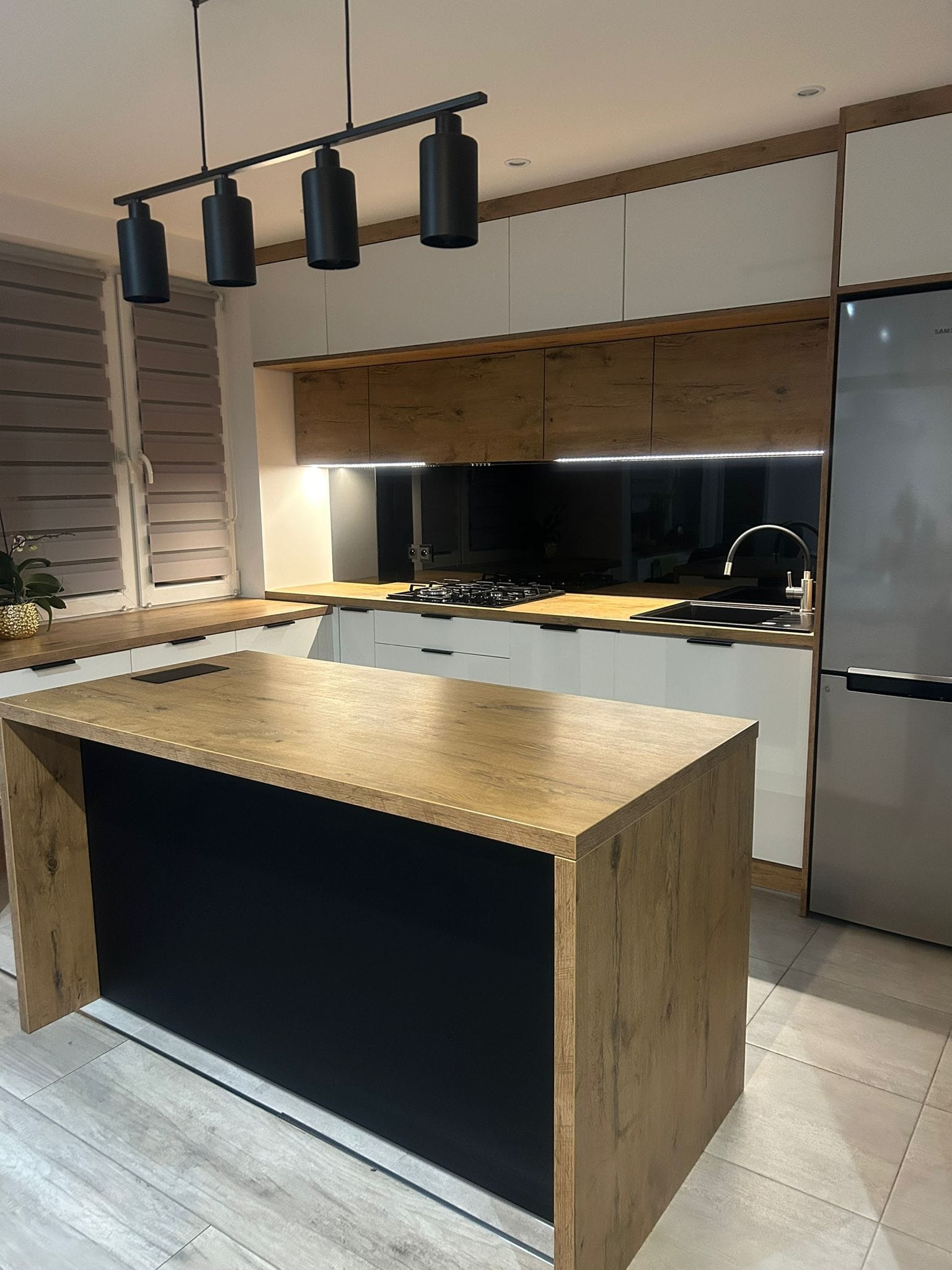 Nowoczesna kuchnia z wyspą: białe i drewniane szafki, czarny panel nad blatem, oświetlenie punktowe i drewnopodobna wyspa z czarnym frontem.