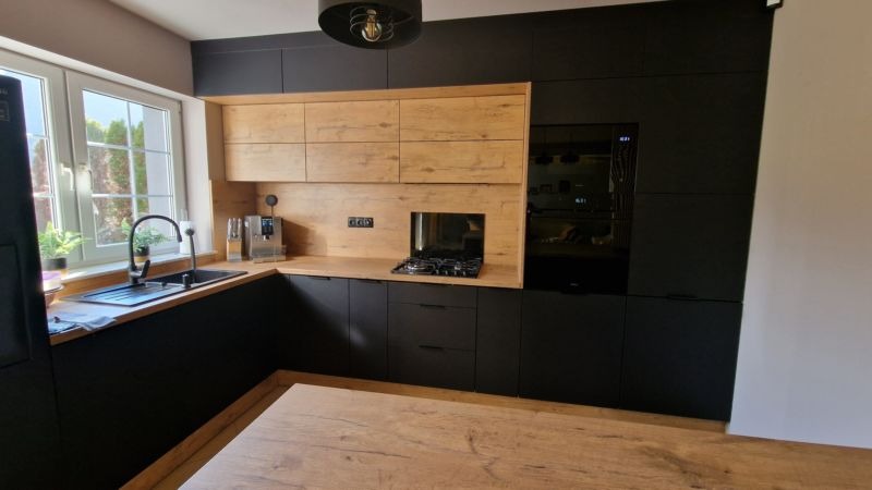 Nowoczesna kuchnia z czarnymi matowymi szafkami dolnymi i drewnopodobnymi górnymi, z wbudowanym piekarnikiem i płytą gazową, widok na okno z zielenią na zewnątrz.