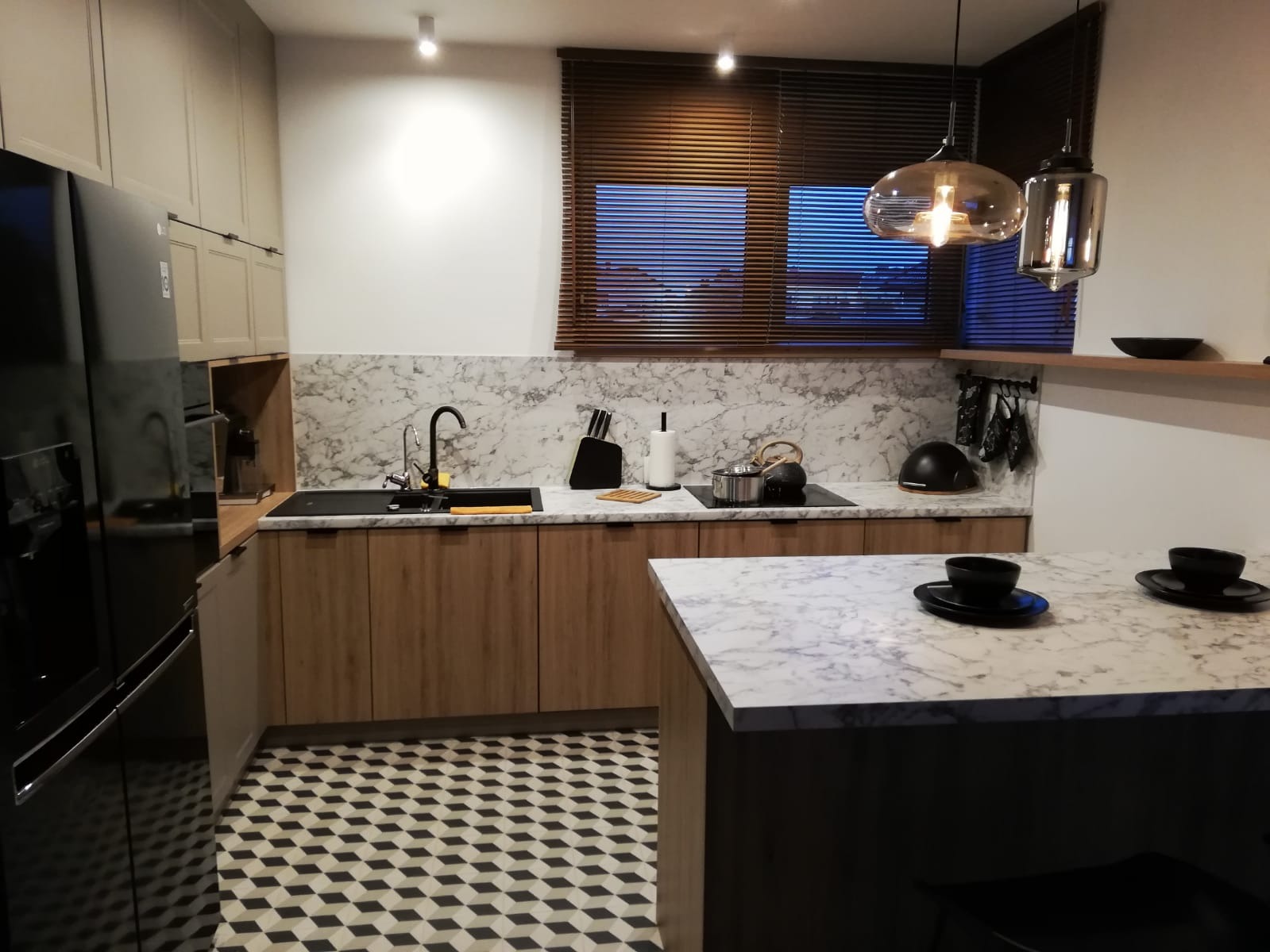 Nowoczesna kuchnia z drewnianymi frontami, marmurowym blatem i geometryczną podłogą w czarno-białe romby; nad wyspą kuchenną wiszą szklane lampy.