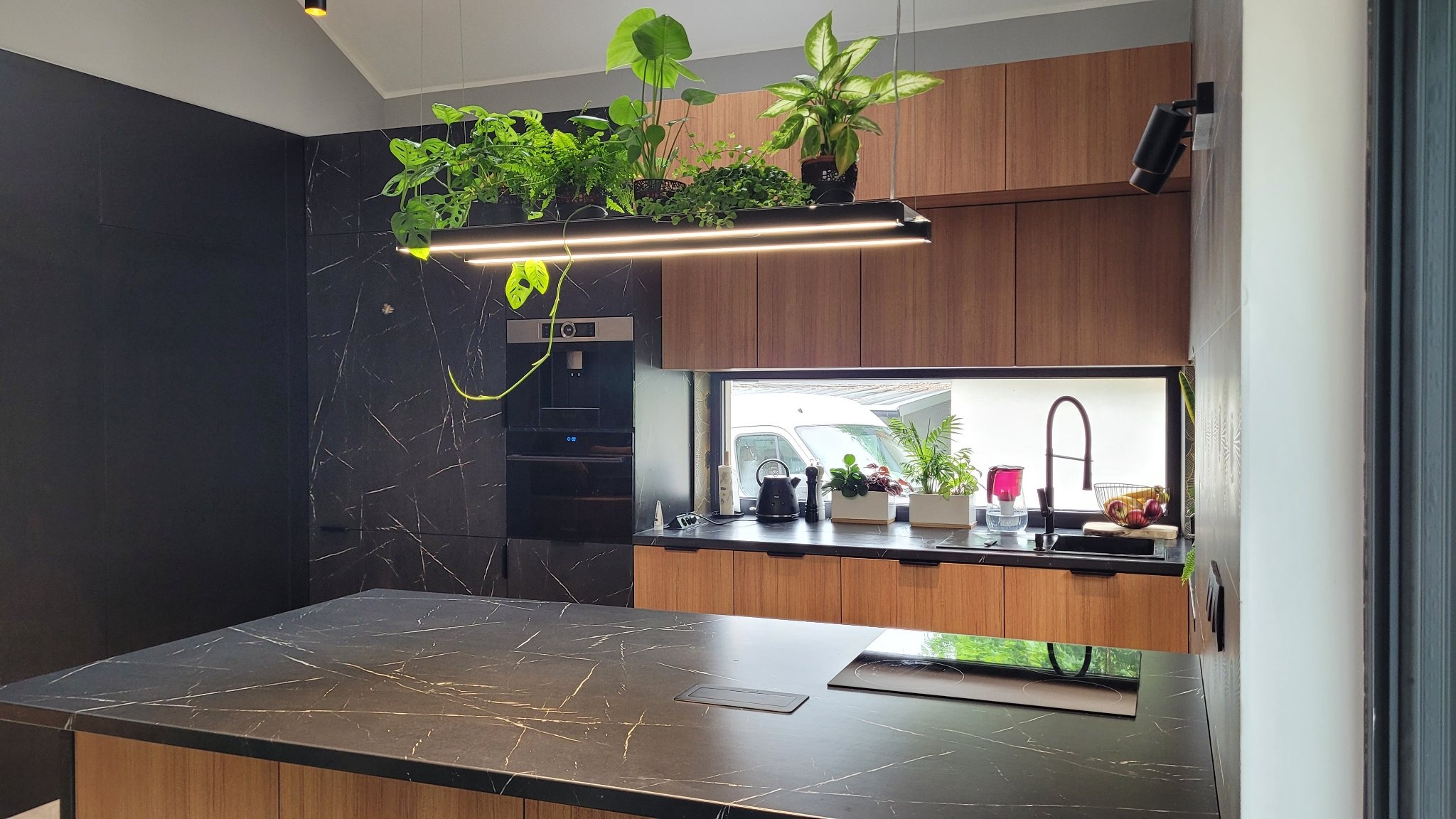 Nowoczesna kuchnia z czarnymi marmurowymi ścianami i drewnianymi szafkami, oświetlona lampą z roślinami, widok na okno z czarnym blatem i zlewem.