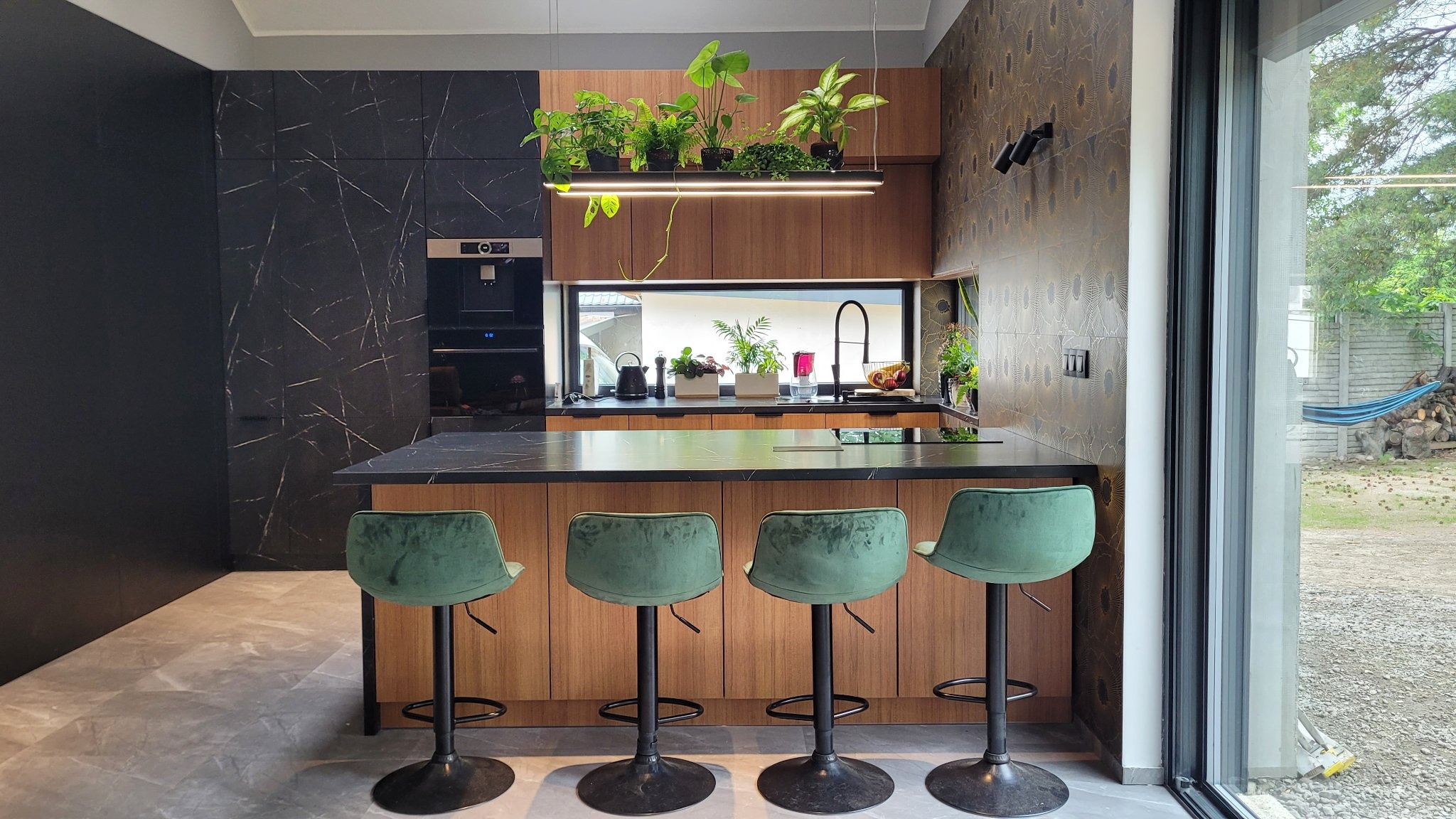 Nowoczesna kuchnia z czarnym blatem, drewnianymi szafkami i wiszącymi roślinami nad wyspą, widok na ogród z hamakiem przez duże okno.
