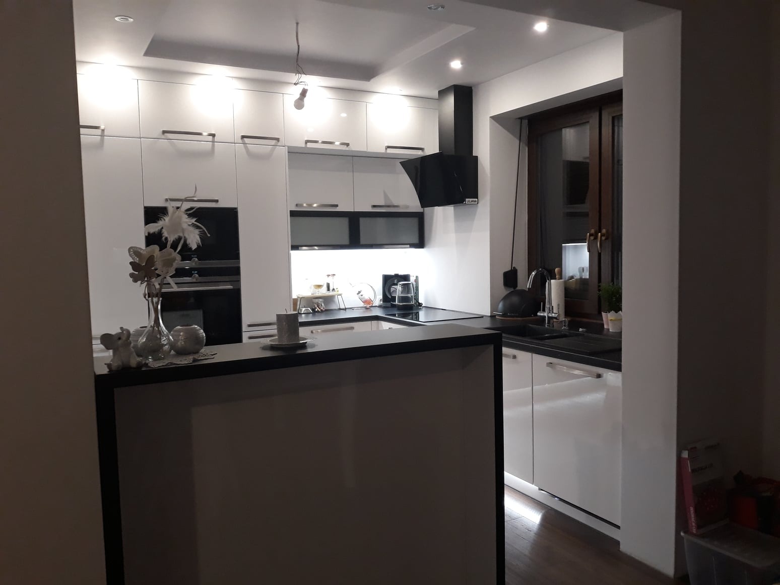 Nowoczesna kuchnia z białymi lakierowanymi frontami, czarnym blatem i wyspą kuchenną z dekoracjami: figurką słonia, wazonem z kwiatami i filiżanką na blacie.