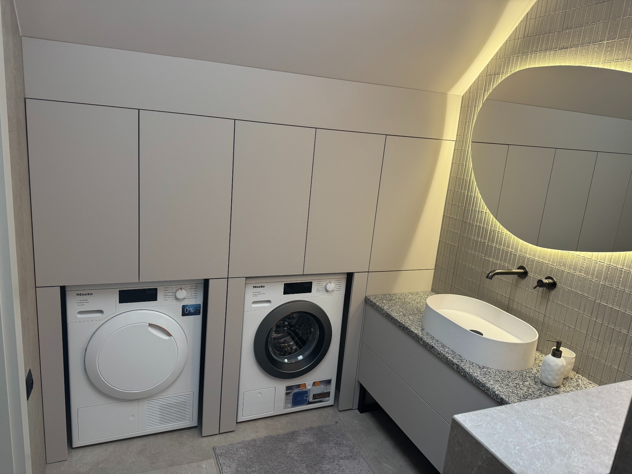 Szara zabudowa meblowa w łazience z pralką i suszarką Miele, owalna umywalka nablatowa na blacie z szarego granitu, okrągłe lustro z podświetleniem LED.
