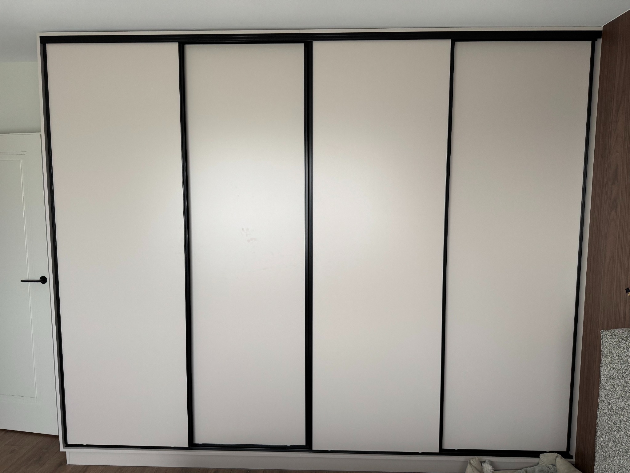 Czterodrzwiowa szafa wnękowa z przesuwnymi, białymi frontami i czarnymi ramami, widoczna z boku drzwi wejściowe i fragment ściany z wykończeniem drewnopodobnym.