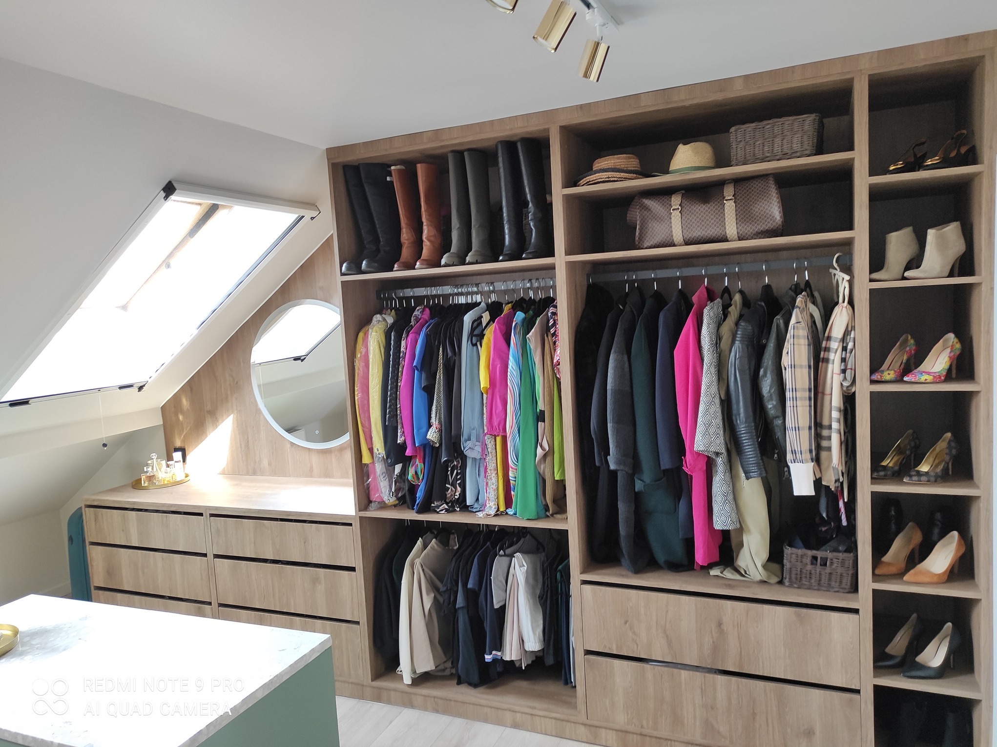 Jasna garderoba na poddaszu ze skośnym oknem, zorganizowana przestrzeń z butami, ubraniami i akcesoriami na półkach i wieszakach, uzupełniona toaletką z okrągłym lustrem.