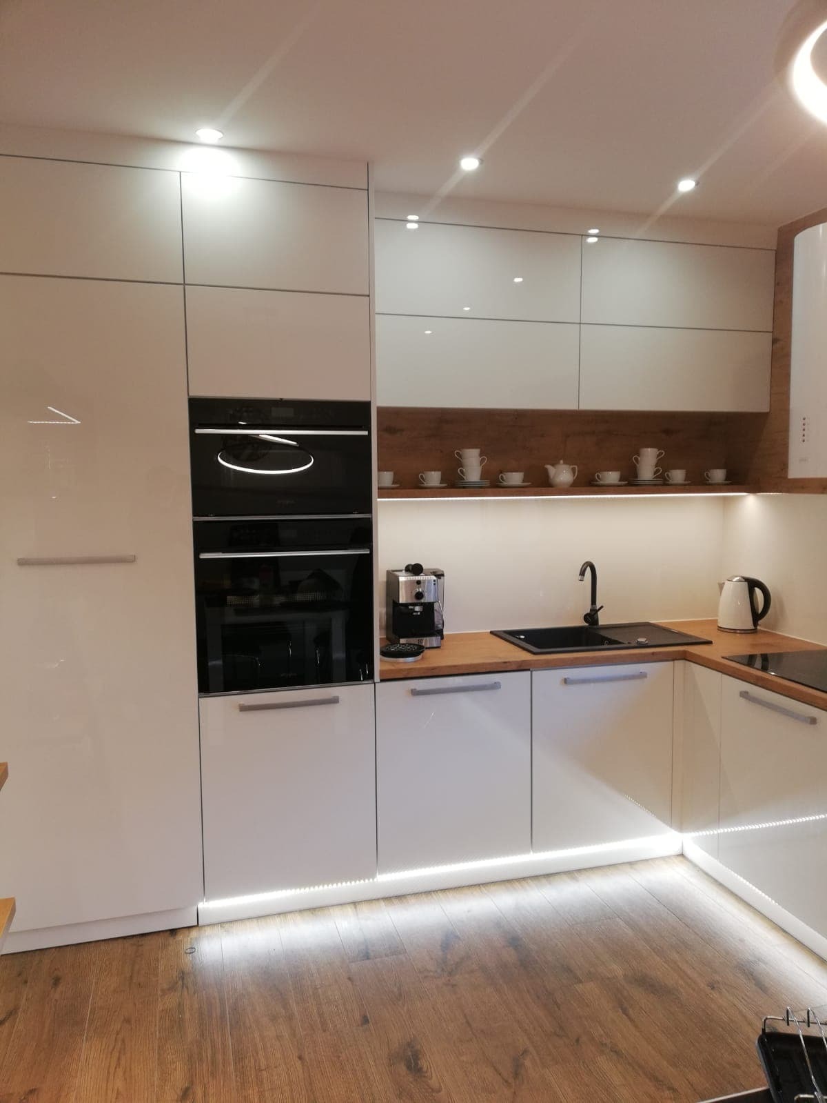 Nowoczesna kuchnia z białymi, lakierowanymi frontami, drewnianym blatem i podświetleniem LED w dolnej części szafek. Na półce dekoracyjne filiżanki i imbryk.