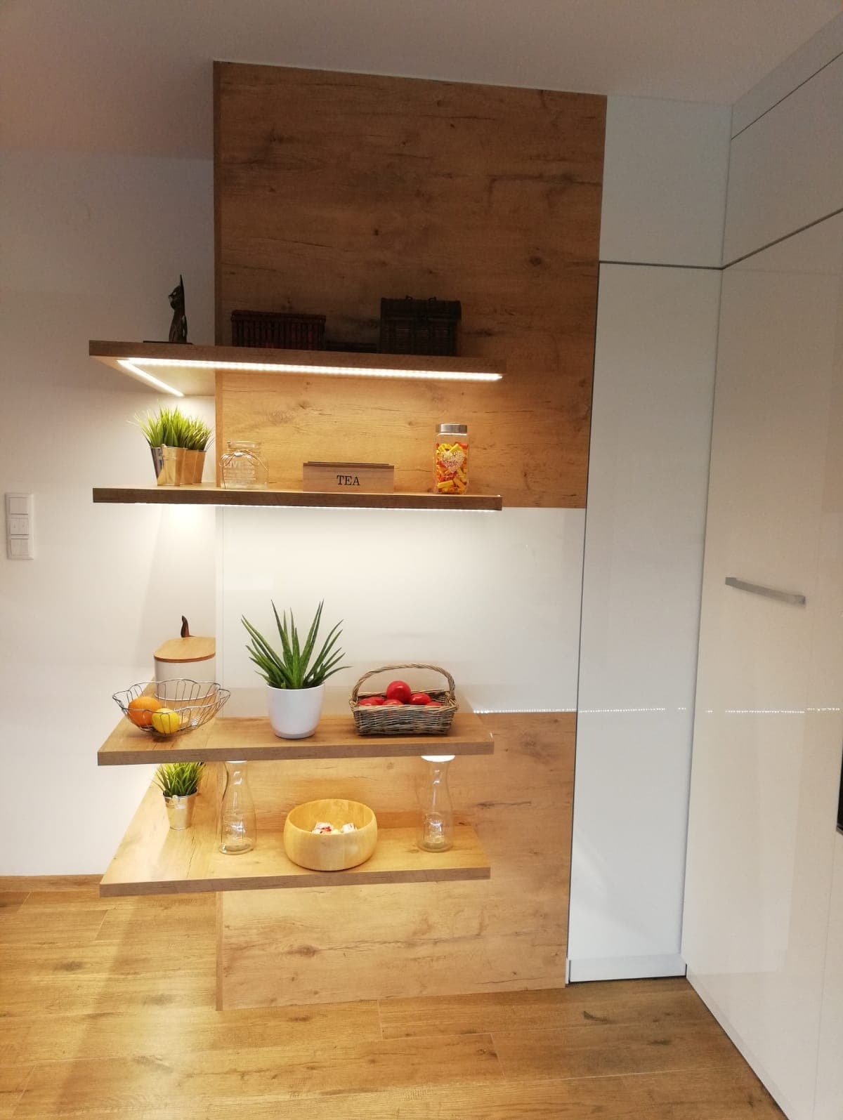 Półki z podświetleniem LED w nowoczesnej kuchni, dekoracyjne przedmioty i produkty spożywcze na drewnianych półkach, białe fronty szafek w tle.