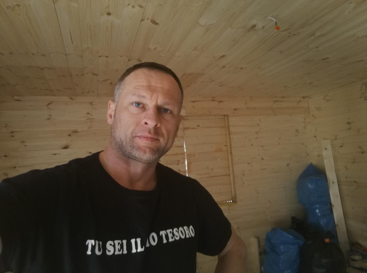 Mężczyzna w czarnej koszulce z napisem 'TU SEI IL MIO TESORO' pozuje we wnętrzu wykończonym boazerią, widoczne niebieskie worki i wystający kabel elektryczny.