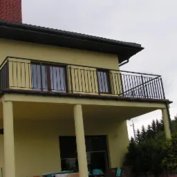 Żółty dom z balkonem i czarną, prostą barierką. Widok z dołu, na drugim planie drzewa i pochmurne niebo. Minimalistyczny design.