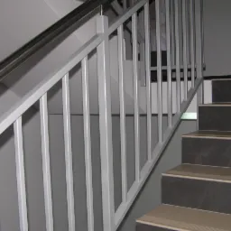 Nowoczesne schody z szarymi stopniami i białą balustradą z metalowymi, pionowymi prętami oraz stalową poręczą. Minimalistyczny design w jasnym wnętrzu.
